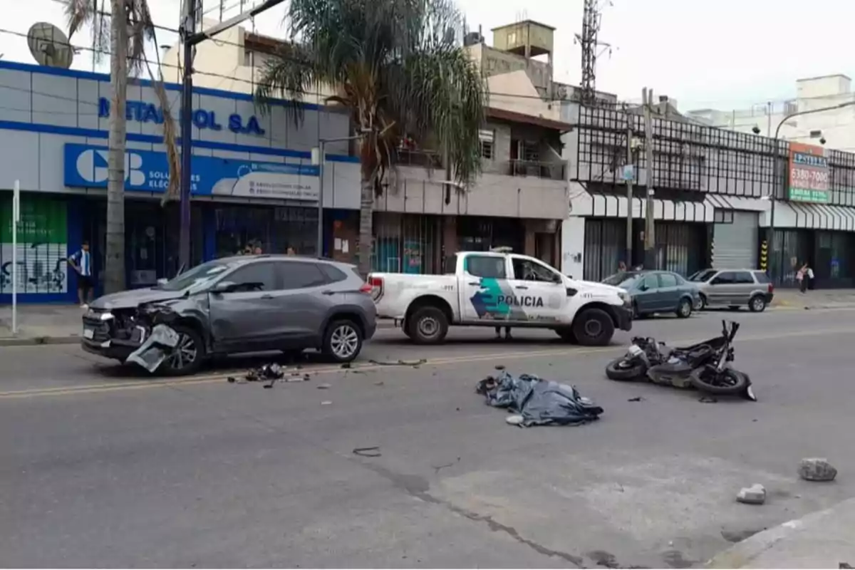 Accidente de tráfico en una calle con un coche gris dañado, una motocicleta caída y una camioneta de policía estacionada cerca.