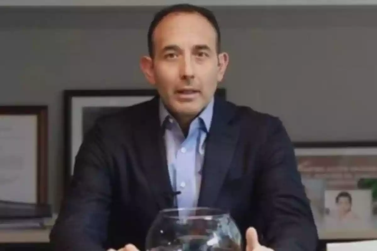 A man in a dark suit and blue jersey is sitting in front of a table with a glass container.