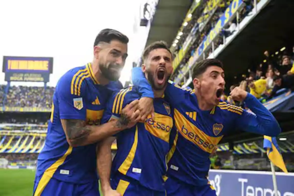 Tres jugadores de fútbol con uniformes azules y amarillos celebran efusivamente en un estadio lleno de aficionados.