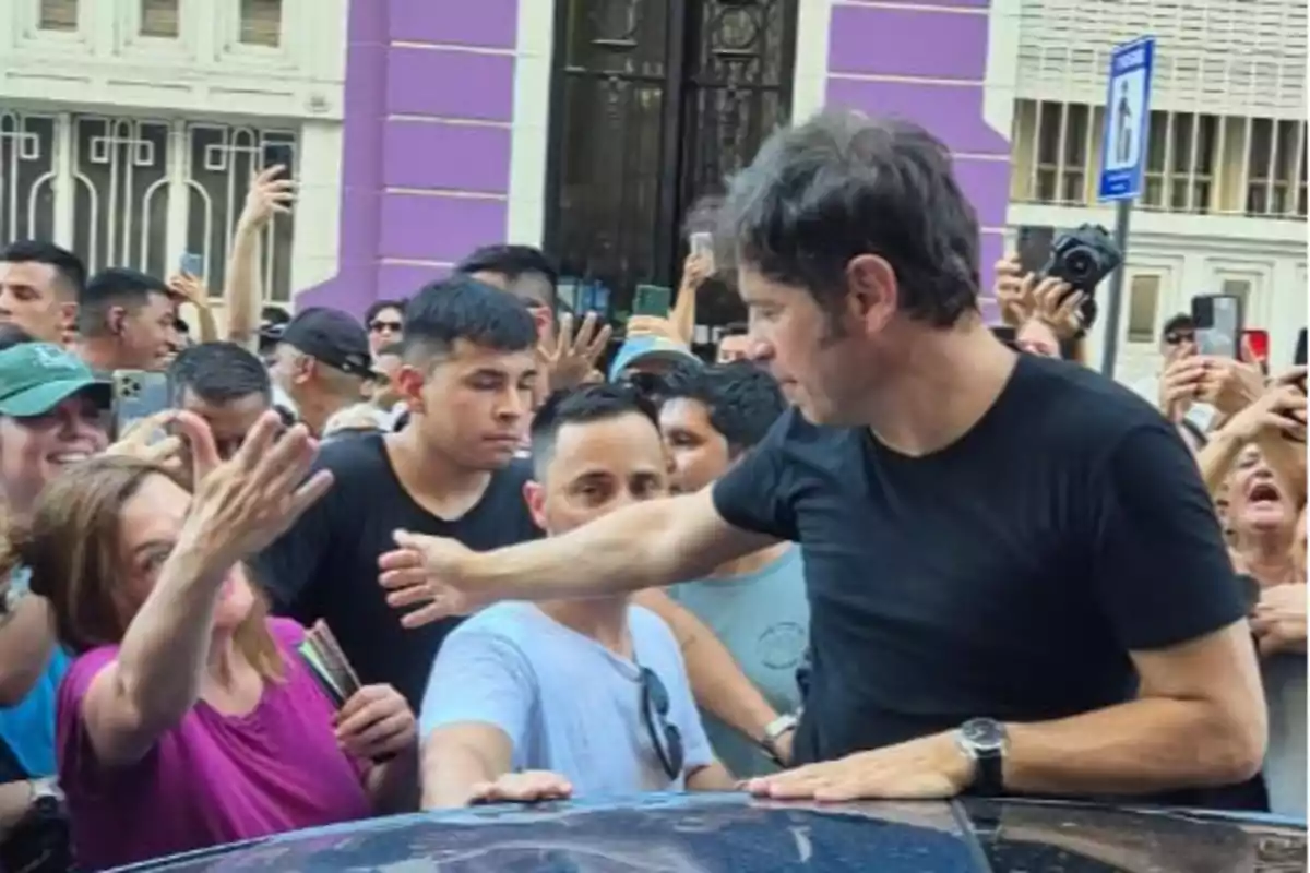 Un hombre de camiseta negra saluda a una multitud de personas que lo rodean mientras algunos toman fotos con sus teléfonos.