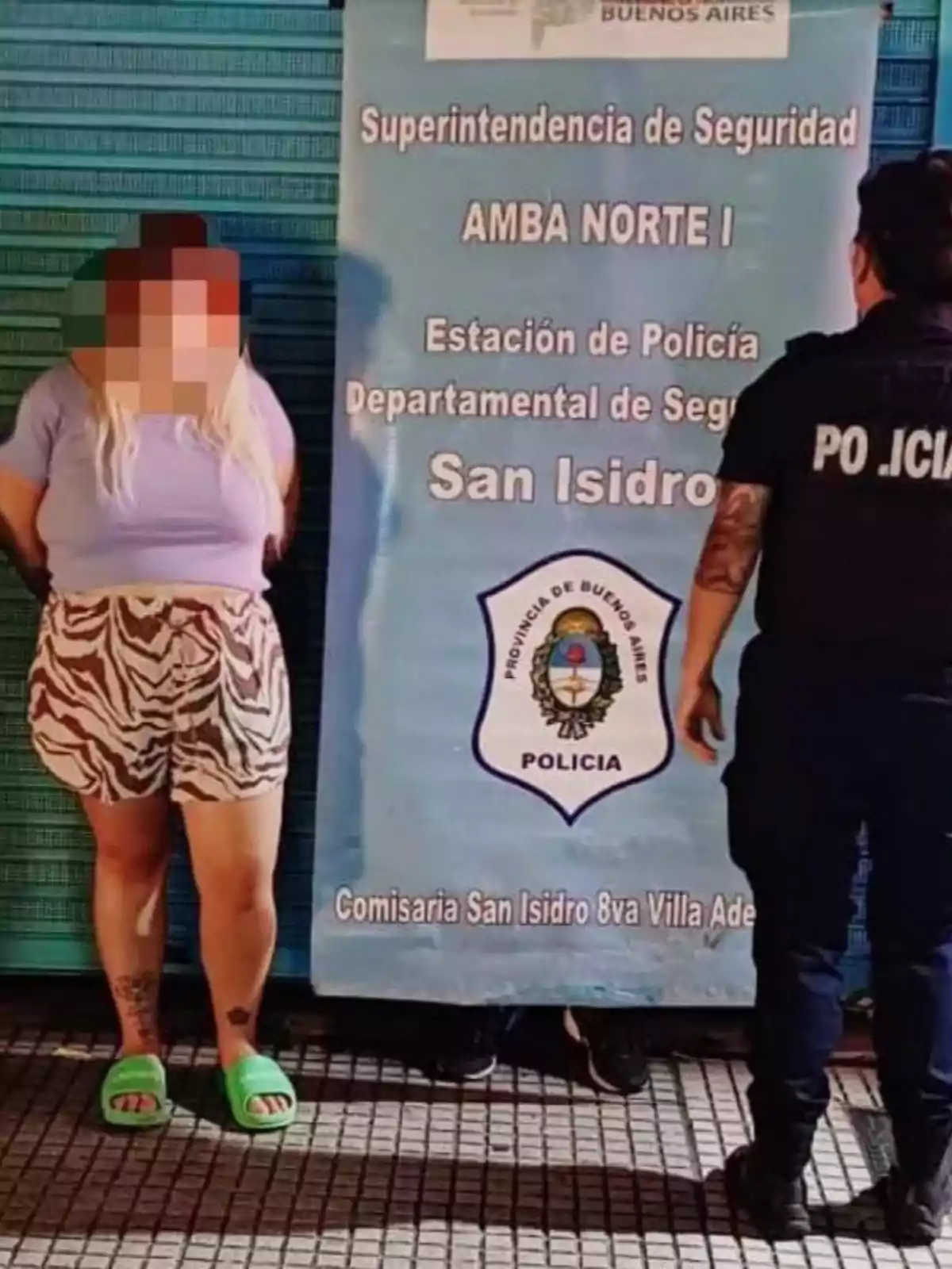 Una persona con el rostro pixelado está de pie junto a un cartel de la policía de San Isidro, acompañada por un oficial.