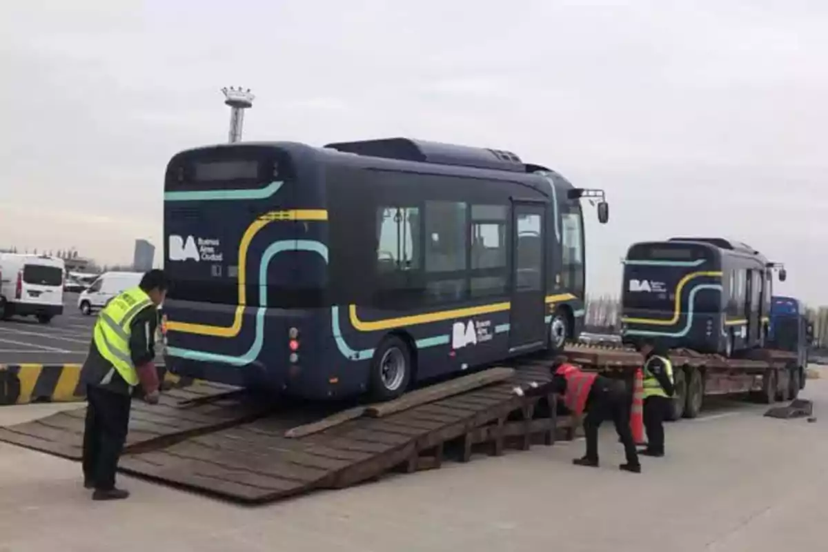 Un autobús eléctrico siendo cargado en un remolque por trabajadores con chalecos reflectantes en un área de estacionamiento.