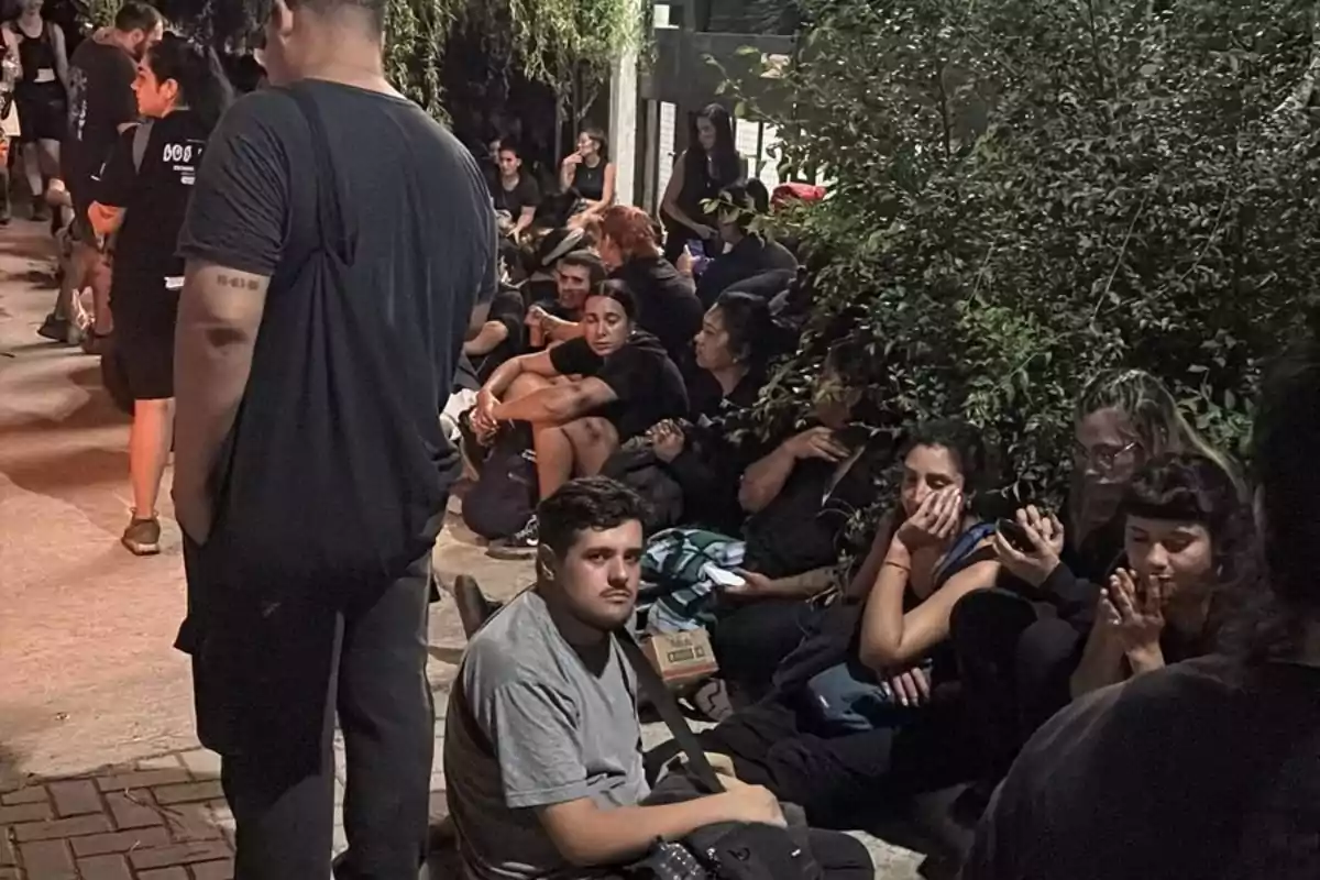 Un grupo de personas sentadas y de pie en una acera, algunas mirando sus teléfonos, rodeadas de vegetación.