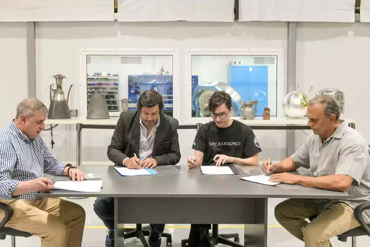 Cuatro personas sentadas en una mesa firmando documentos en un entorno industrial con piezas metálicas en el fondo.