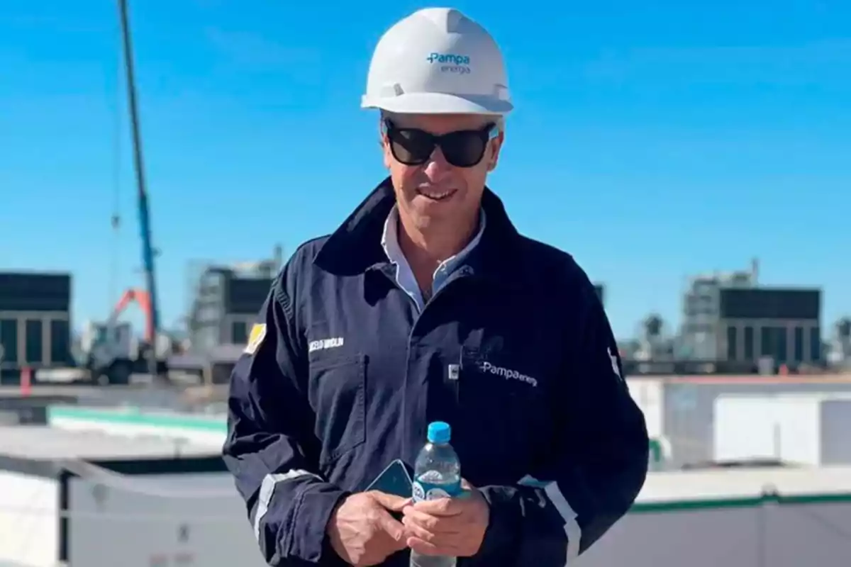 Persona con casco y gafas de sol, vistiendo un uniforme de trabajo de Pampa Energía, sosteniendo una botella de agua y un teléfono móvil, con un fondo de construcción industrial.