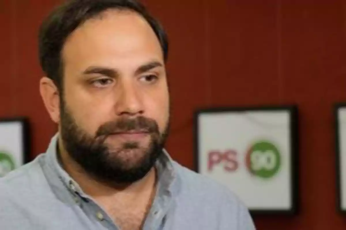 Hombre con barba y camisa azul claro frente a un fondo con carteles borrosos.