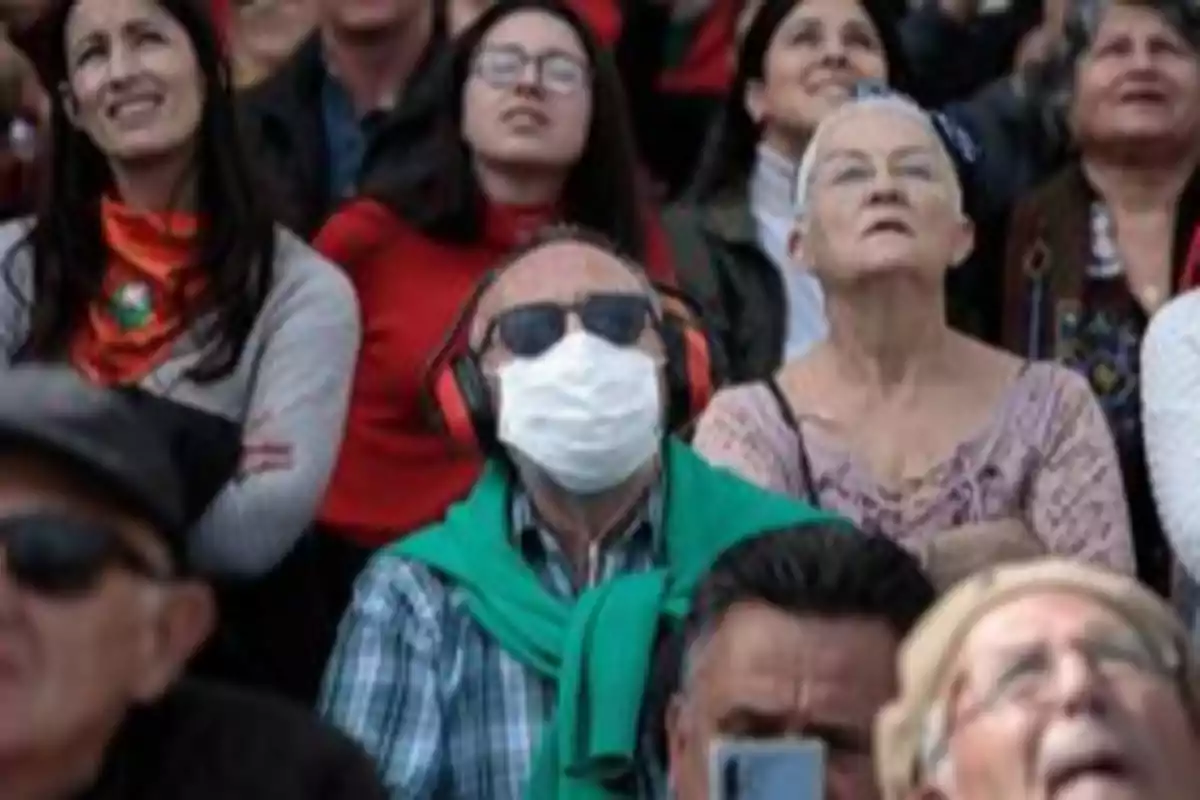 Un grupo de personas observa algo en el cielo, una de ellas lleva mascarilla, gafas de sol y protectores auditivos por el brote de hMPV en China.