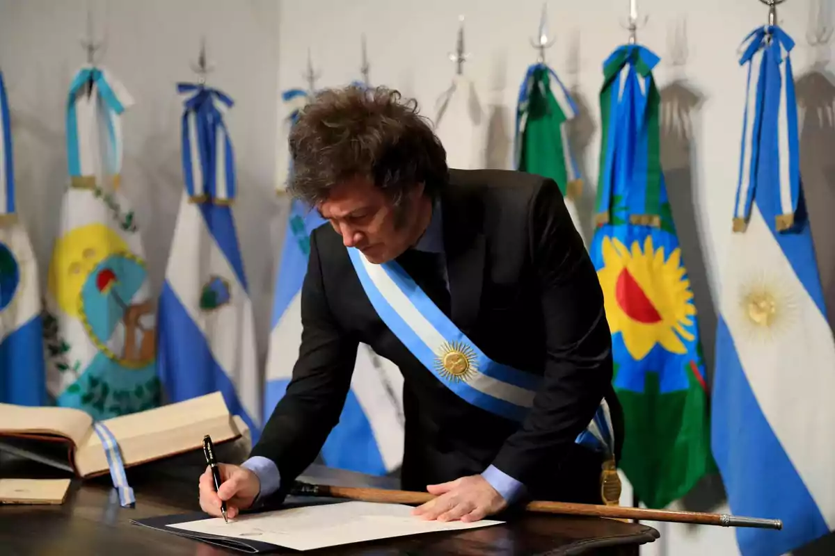 Un hombre con una banda presidencial firma un documento en una mesa con banderas argentinas de fondo.