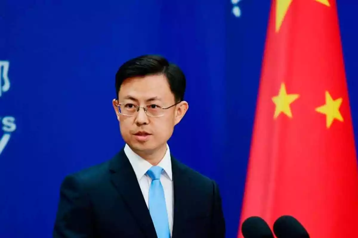 Un hombre con gafas y traje oscuro habla frente a un micrófono con una bandera de China al fondo.