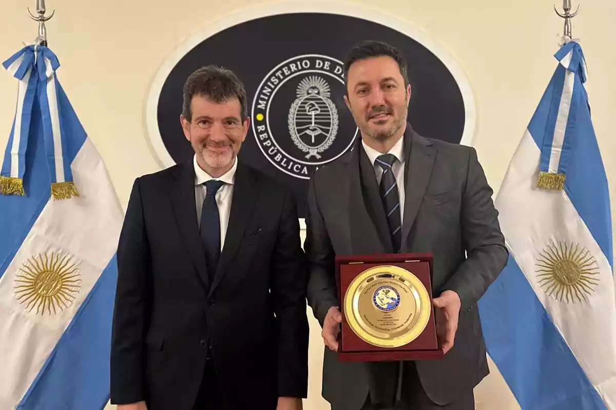 Dos hombres de traje posan frente a un emblema del Ministerio de Defensa de la República Argentina, uno de ellos sostiene una placa dorada enmarcada, con dos banderas argentinas a los lados.