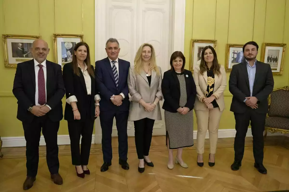 Un grupo de siete personas posando en una habitación con paredes verdes y suelo de madera, con cuadros enmarcados en la pared y una puerta blanca al fondo.