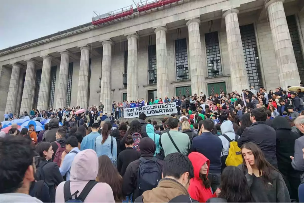 Una multitud se reúne frente a un edificio con columnas, sosteniendo un cartel que dice "Sin presupuesto no hay libertad".