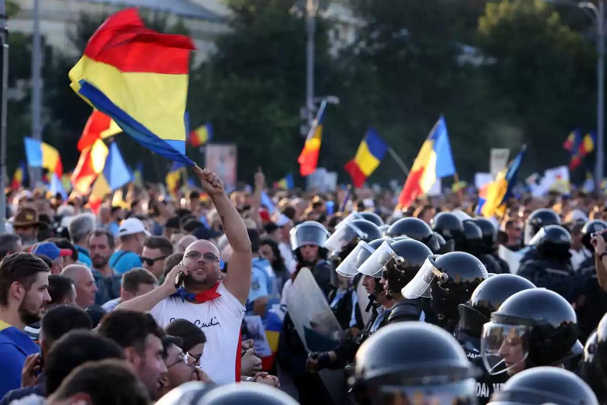 Una multitud de personas se manifiesta en la calle, ondeando banderas de colores rojo, amarillo y azul, mientras un grupo de policías con cascos y escudos observa.