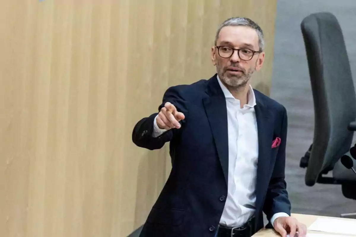 Un hombre con gafas y barba, vestido con traje oscuro y camisa blanca, señala con el dedo mientras está de pie junto a una mesa en un entorno de oficina o conferencia.