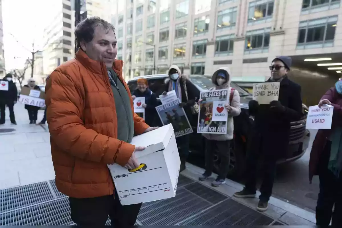 Un hombre con chaqueta naranja lleva una caja mientras un grupo de personas sostiene carteles de agradecimiento en una calle urbana.