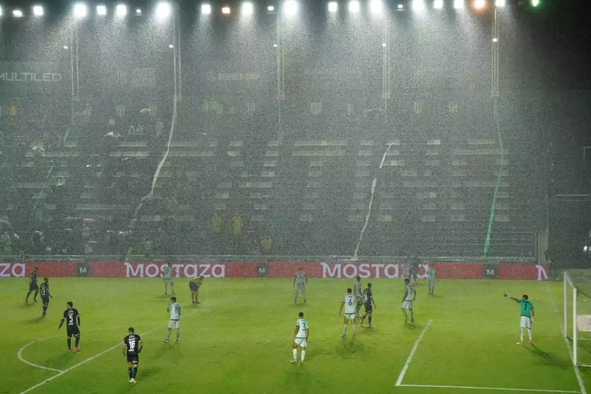 Jugadores de fútbol en un campo bajo la lluvia intensa durante un partido nocturno con luces brillantes iluminando el estadio y gradas parcialmente vacías.