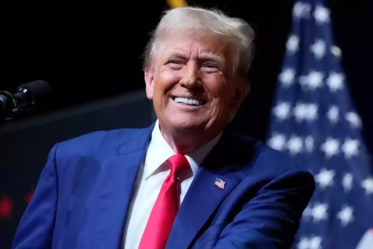 Un hombre sonriente con traje azul y corbata roja frente a una bandera de Estados Unidos.