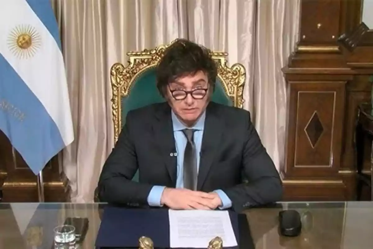 Un hombre con gafas y traje oscuro está sentado en un escritorio con una bandera de Argentina al fondo.