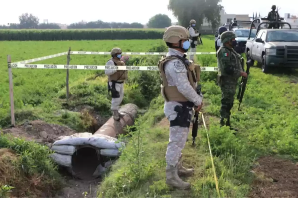 Soldados con equipo táctico vigilan un área acordonada con cinta de precaución en un campo verde.