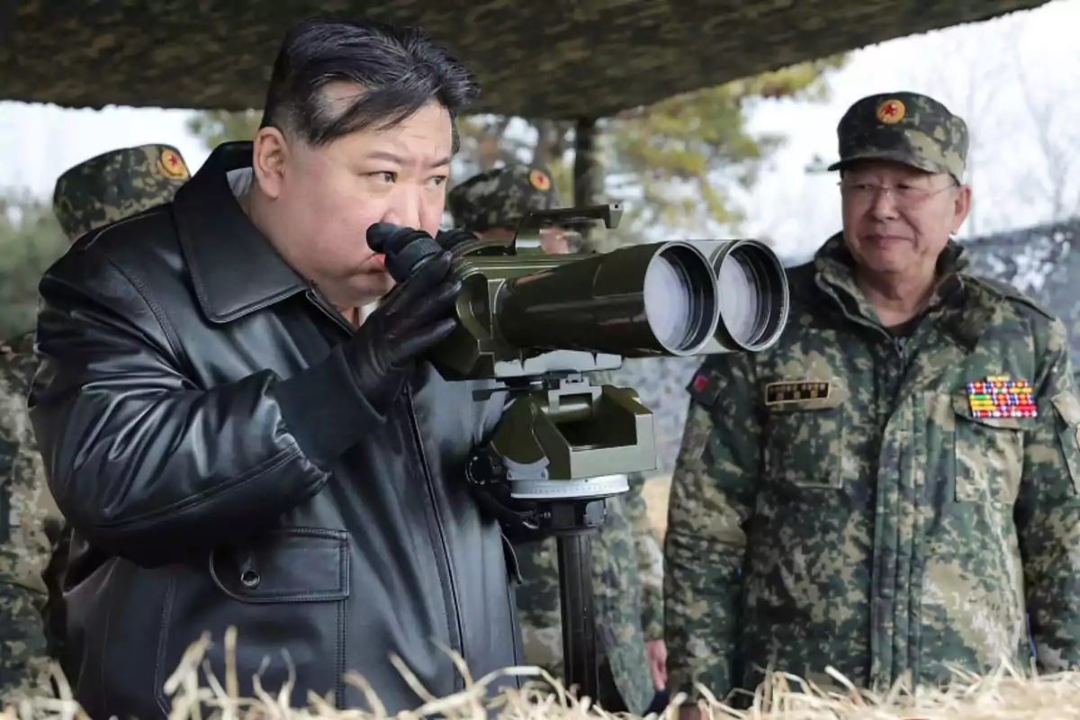 Un hombre con abrigo de cuero negro observa a través de un gran binocular mientras otro hombre con uniforme militar lo observa.