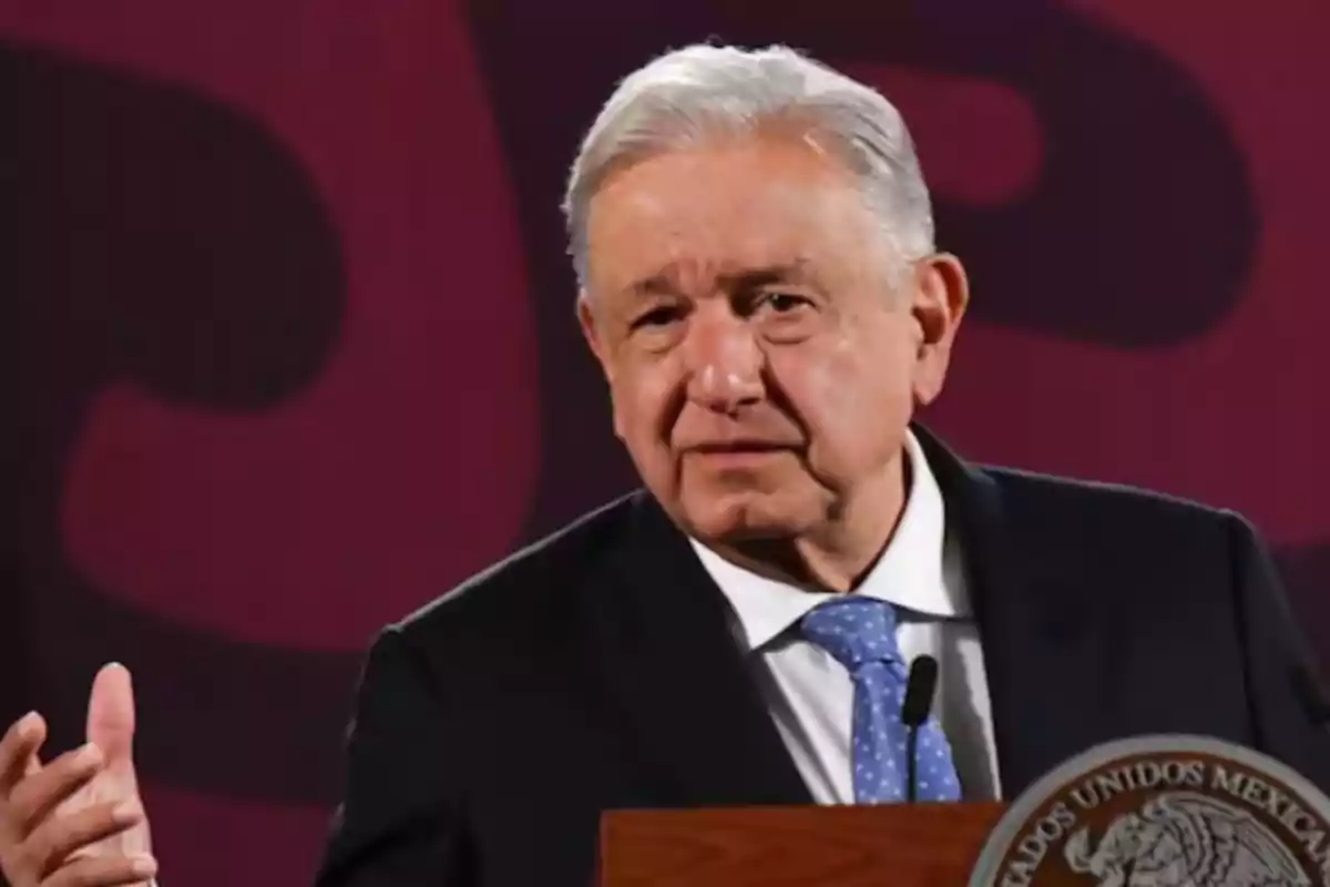 A gray-haired man in a dark suit with a blue tie speaking at a podium with a red background.