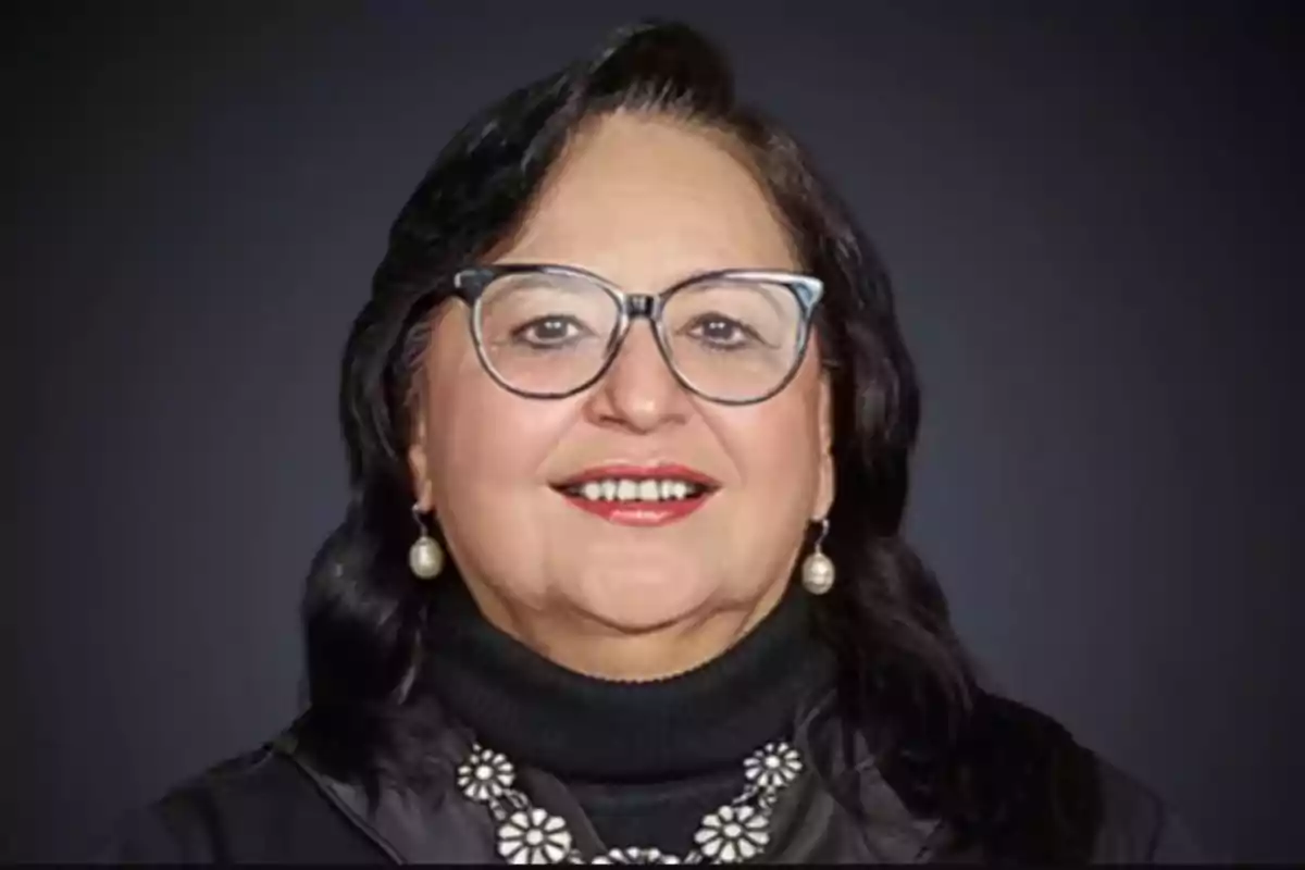 A person with glasses and dark hair smiles in front of a gray background.