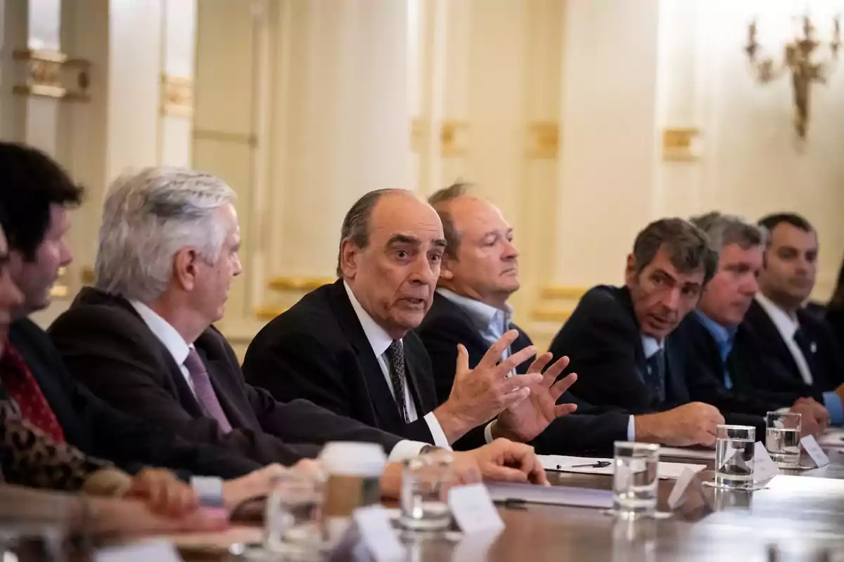 Personas en una reunión formal sentadas alrededor de una mesa, con un hombre mayor hablando y gesticulando.