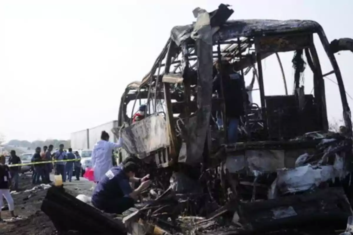 A group of people inspects the charred remains of a bus on a road.