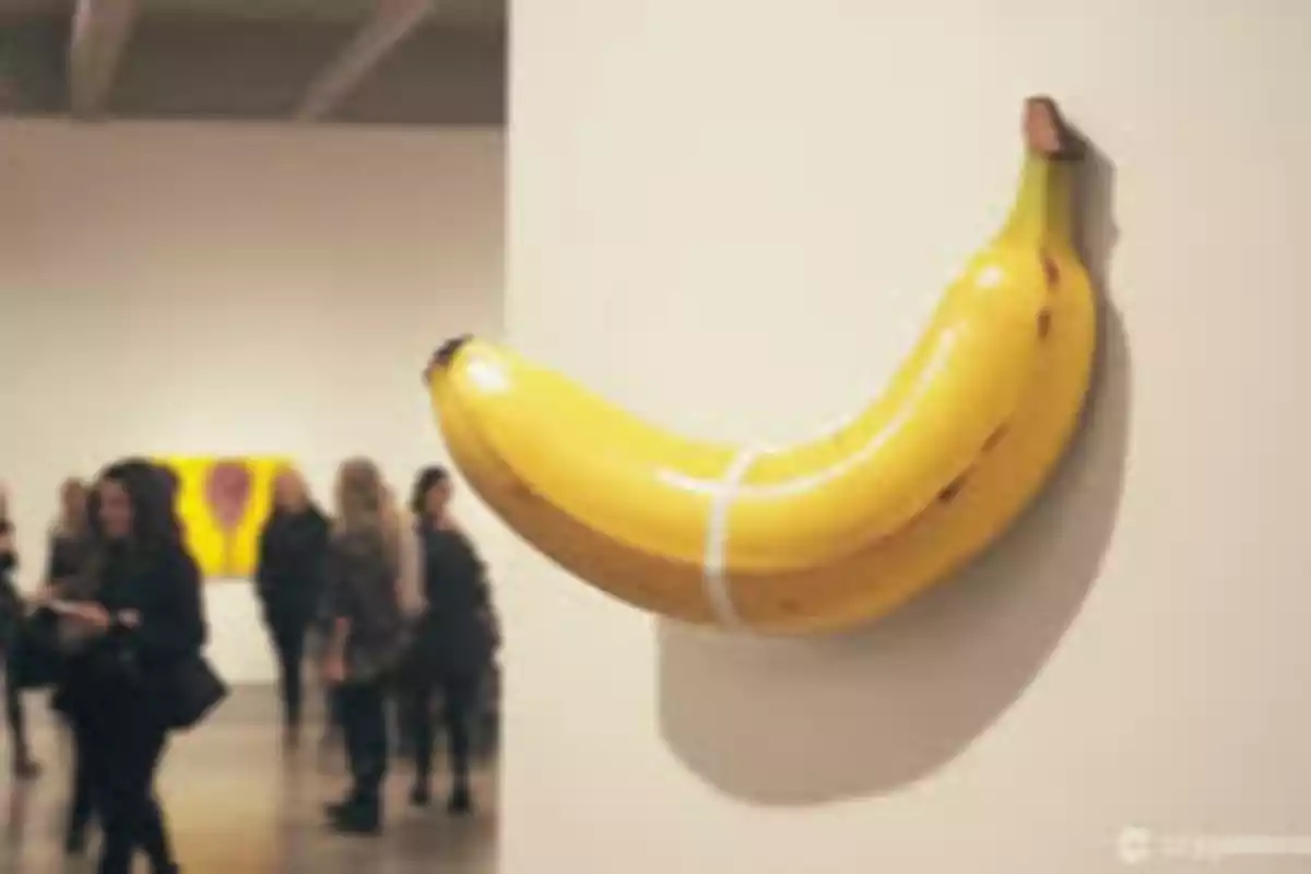 Una banana inflable está pegada a la pared en una galería de arte con personas observando.