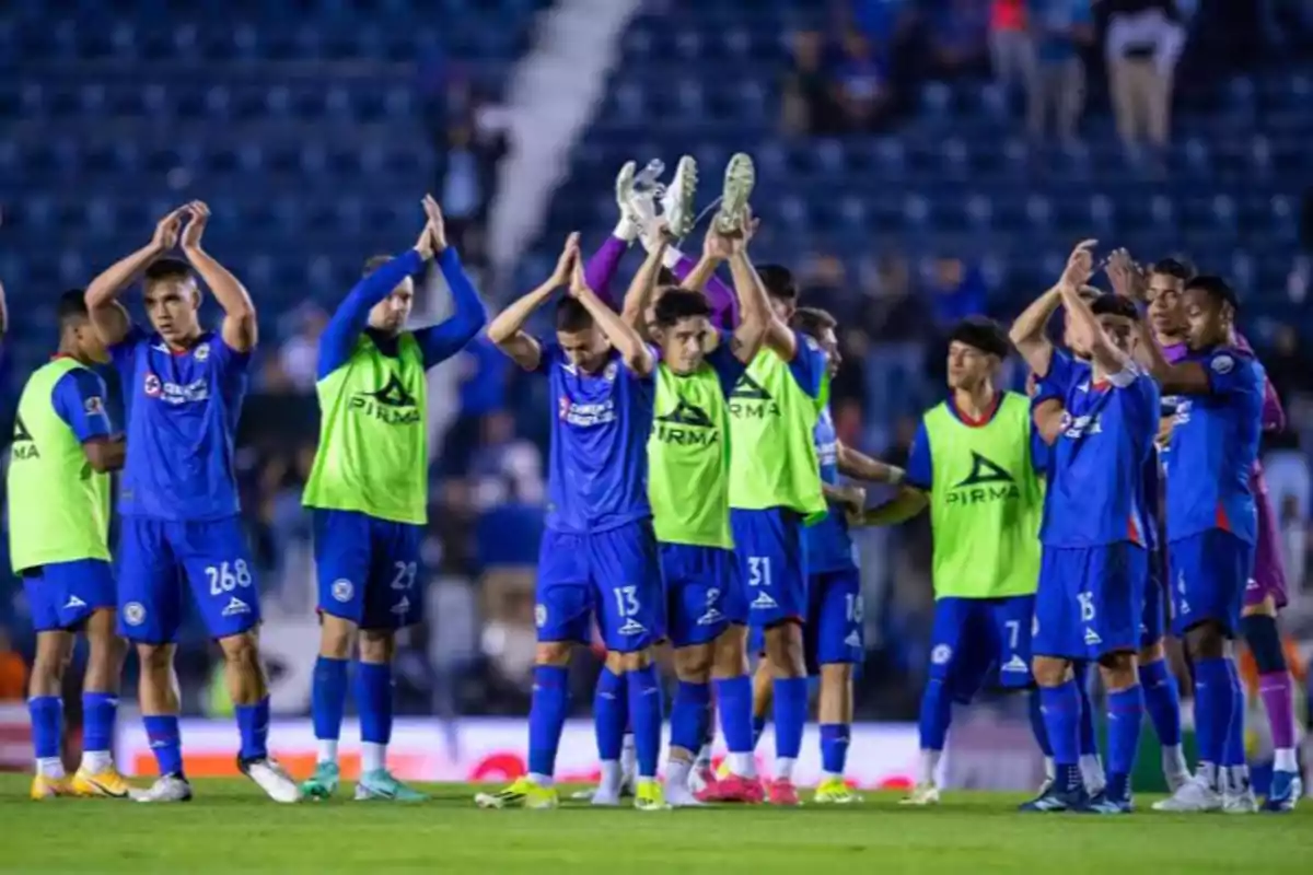 Jugadores de fútbol con uniforme azul y chalecos verdes aplauden en un estadio.