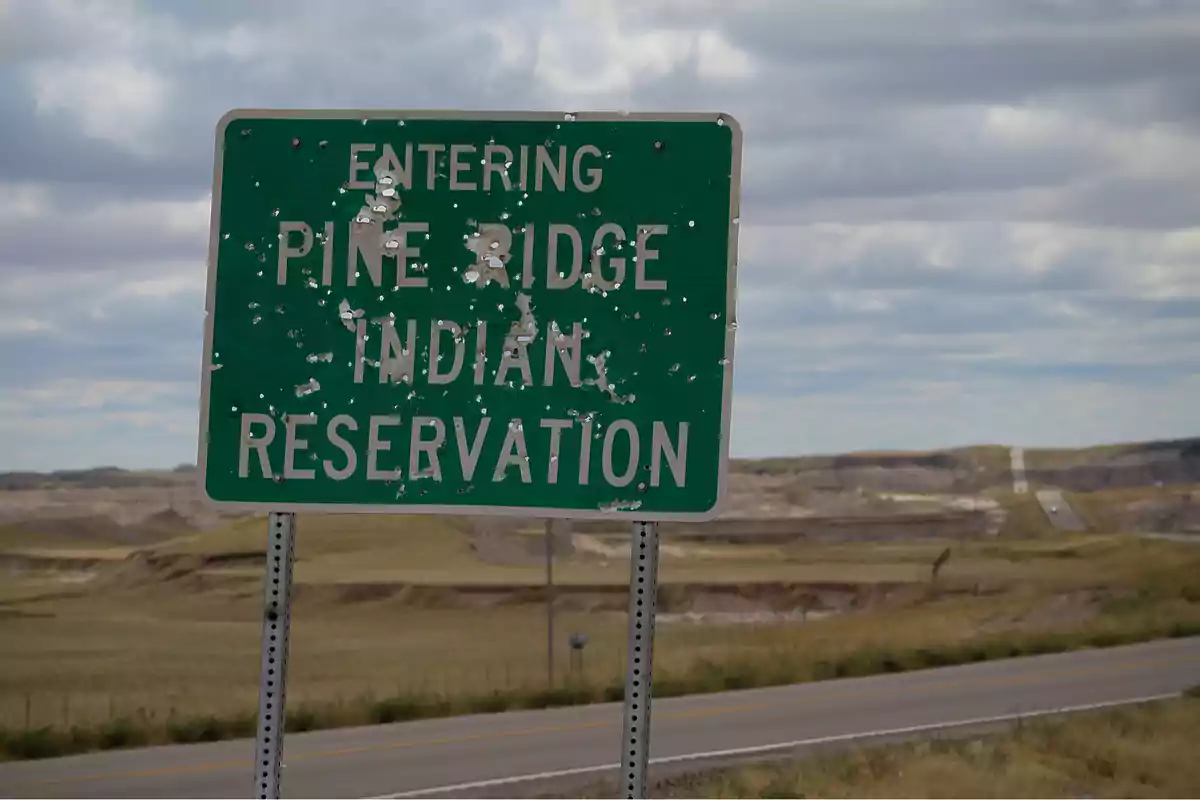 Un cartel verde con texto blanco que indica la entrada a una reserva indígena, con marcas visibles de impactos, en un paisaje de carretera y colinas bajo un cielo nublado.