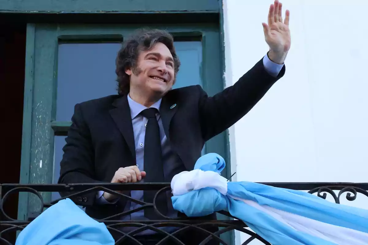 Un hombre sonriente con traje oscuro saluda desde un balcón decorado con telas azules y blancas.