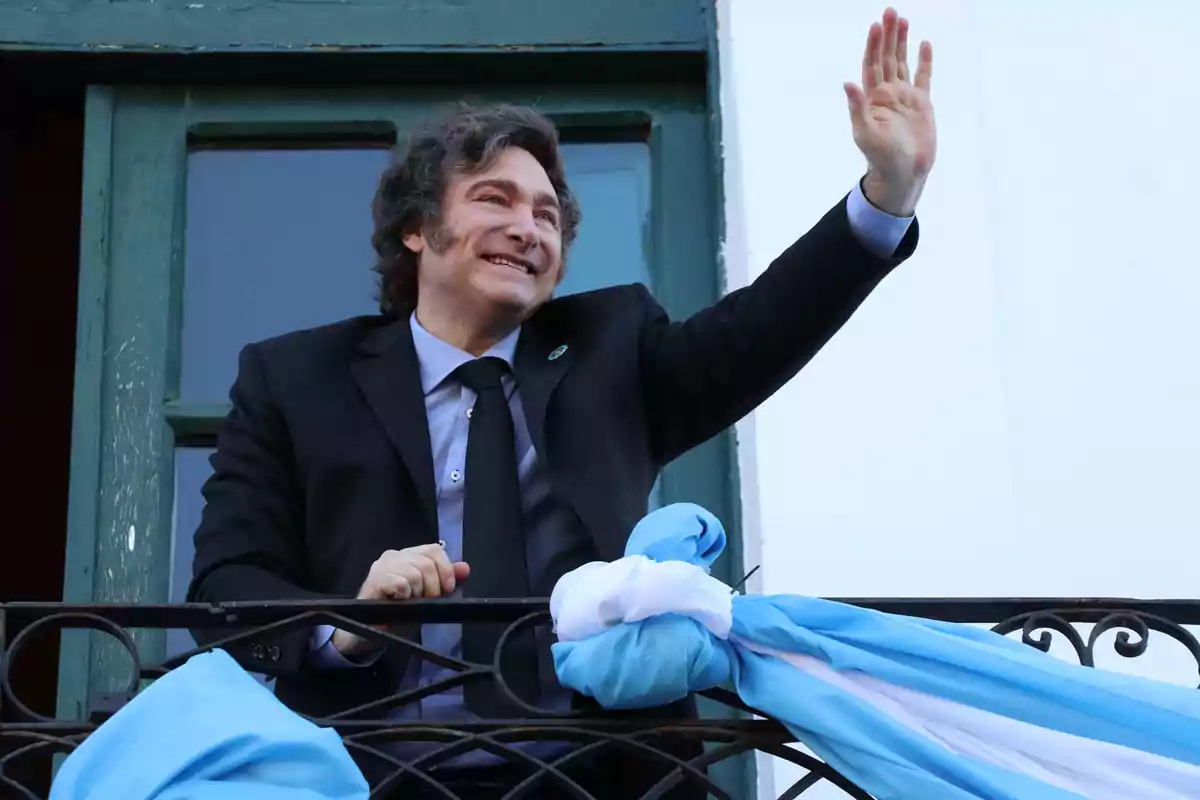 Un hombre sonriente con traje oscuro y corbata saluda desde un balcón decorado con una bandera azul y blanca.