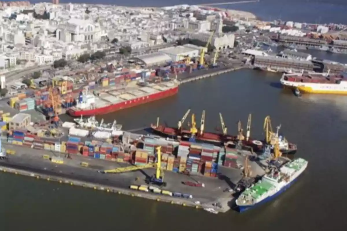 Vista aérea de un puerto con barcos de carga y contenedores en el muelle, rodeado de edificios y el mar al fondo.