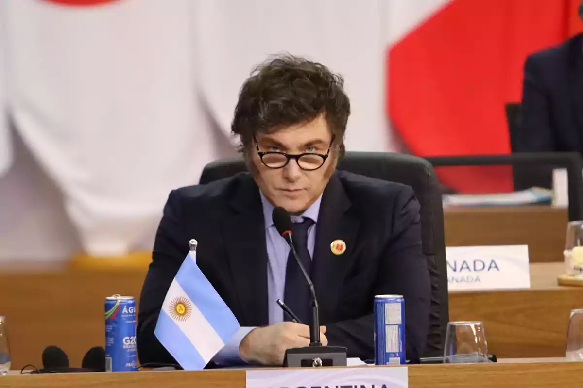 Un hombre con gafas y patillas está sentado en una mesa de conferencia con un micrófono frente a él y una bandera de Argentina.