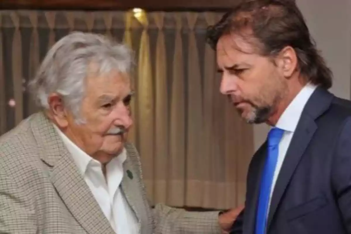 Dos hombres conversando en una habitación con cortinas de fondo.