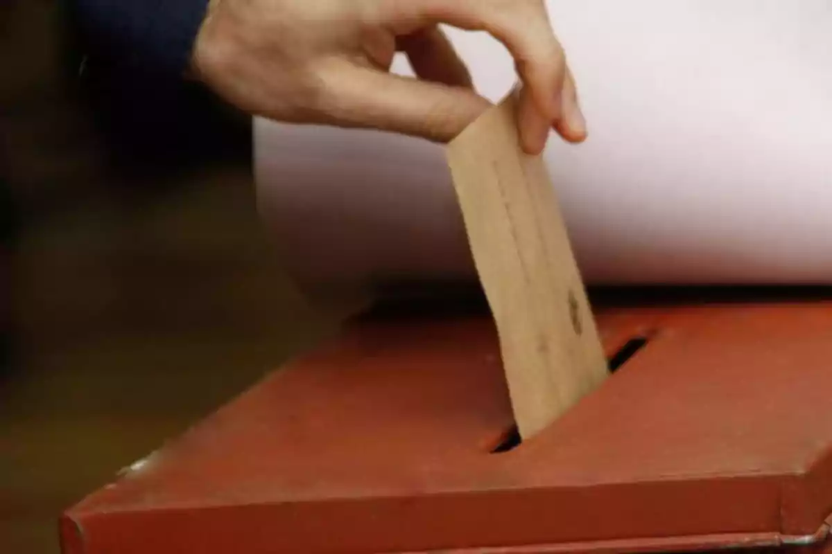 Una mano introduciendo un sobre en una urna de votación.