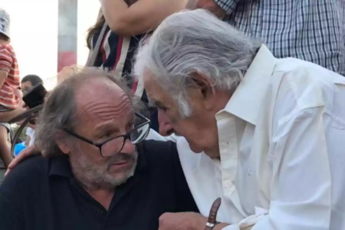 Dos hombres mayores conversan de cerca en un entorno al aire libre, uno lleva gafas y el otro viste una camisa blanca.