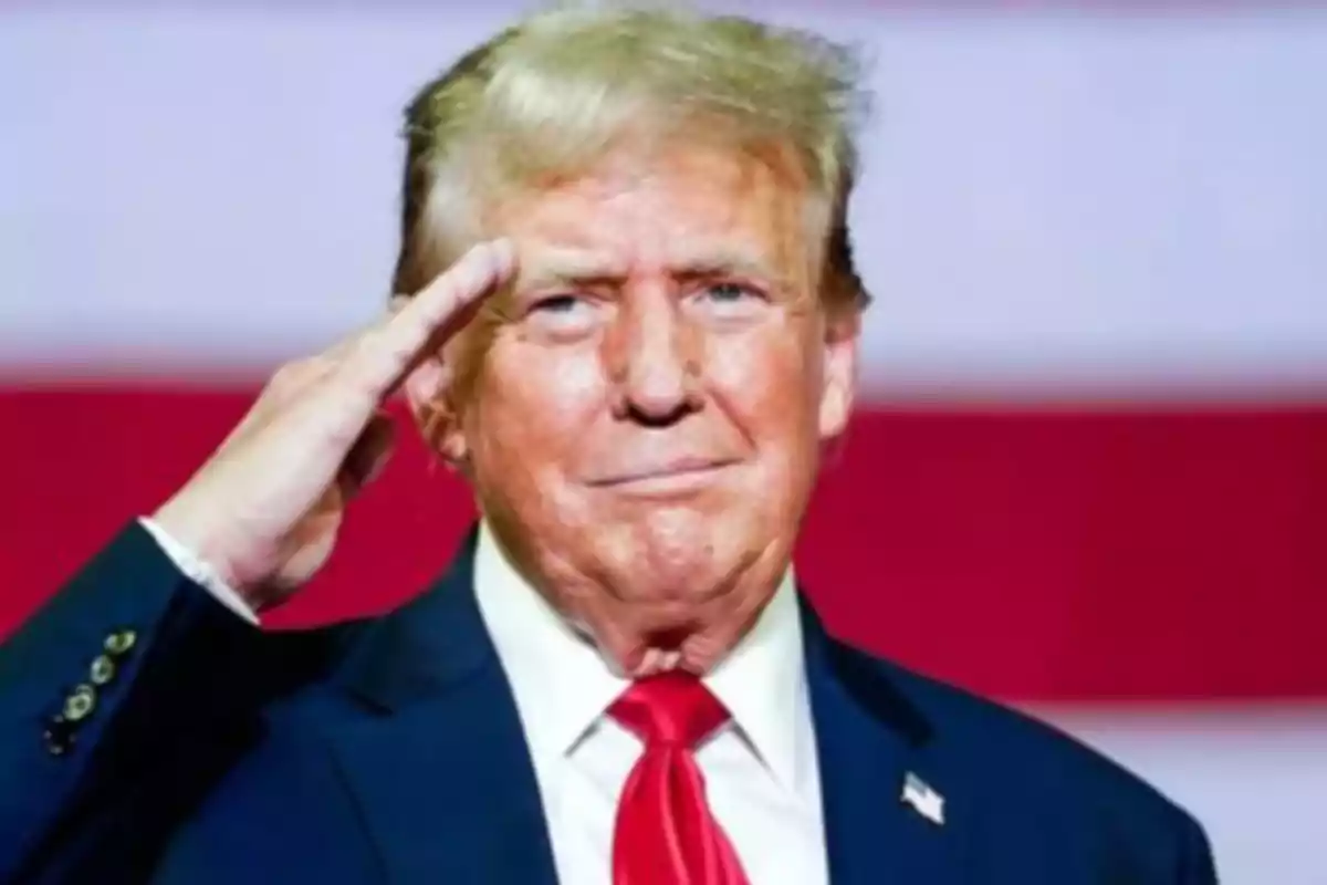 Un hombre de traje azul y corbata roja haciendo un saludo militar frente a un fondo con franjas rojas y blancas.