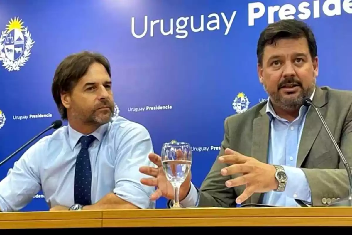 Dos hombres en una conferencia de prensa con un fondo azul que dice 