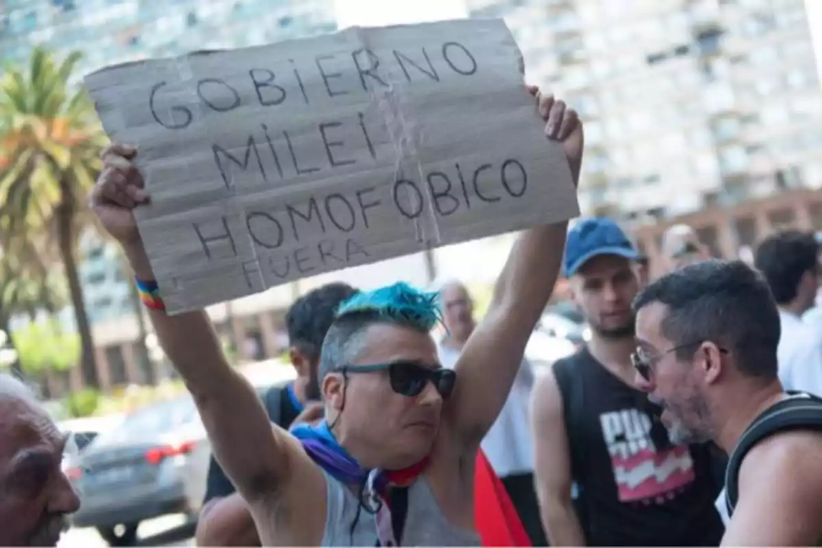 Una persona con cabello azul y gafas de sol sostiene un cartel de protesta en una manifestación al aire libre, rodeada de otras personas.