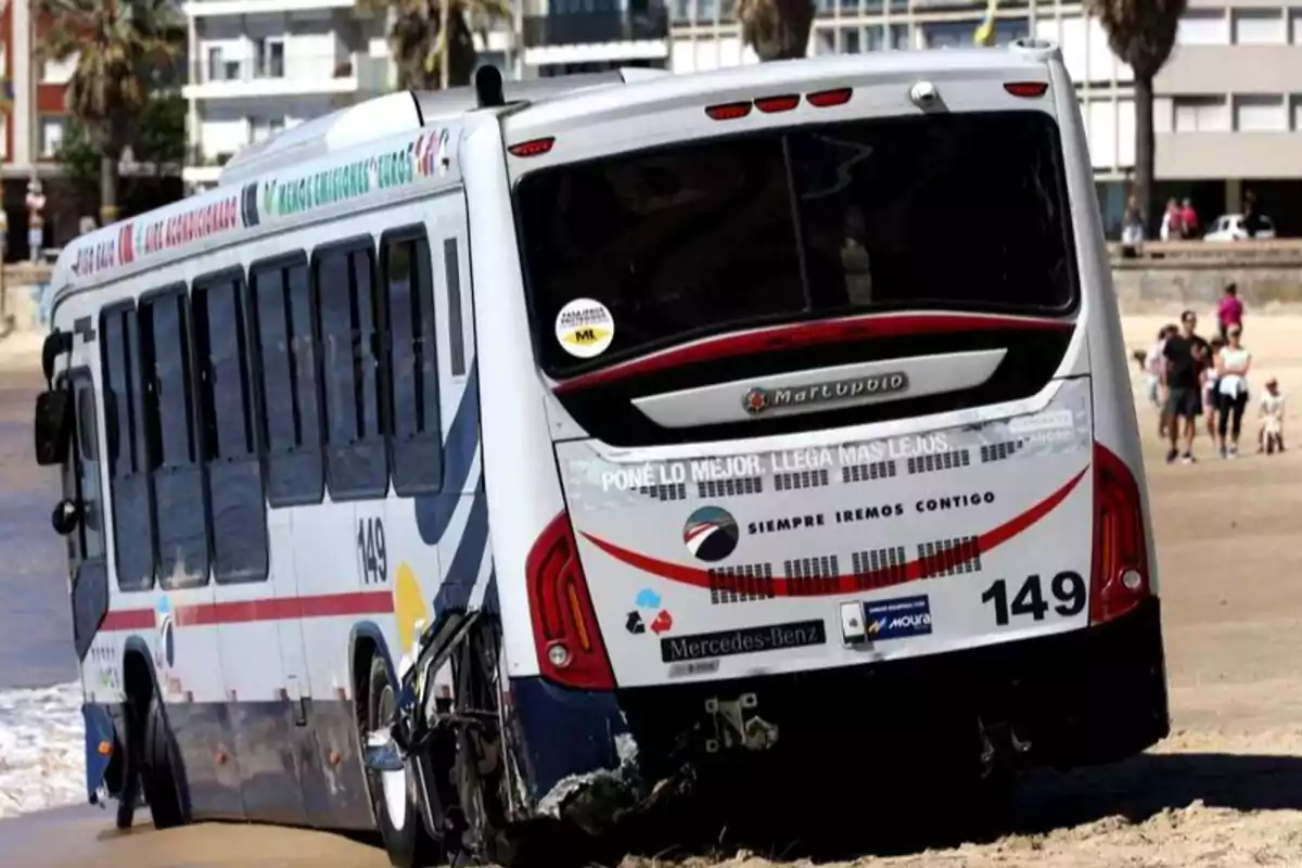 Un autobús atascado en la arena de una playa con personas caminando al fondo.