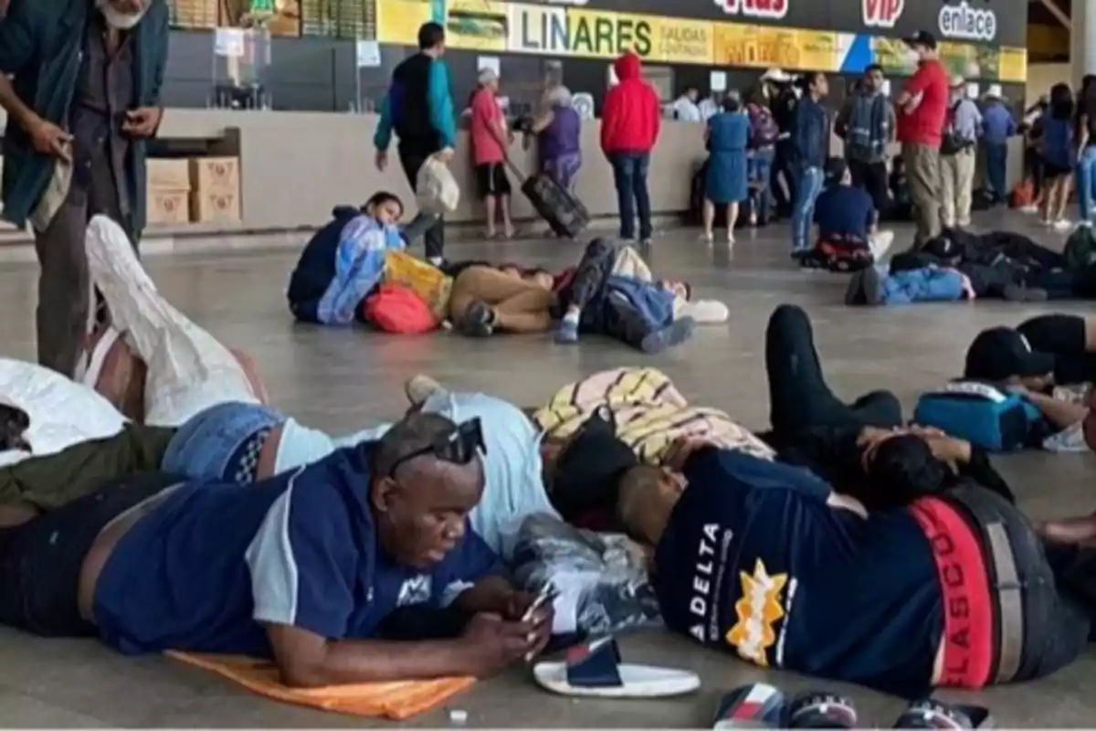 Personas descansando en el suelo de una terminal de autobuses, mientras otras hacen fila en el mostrador.