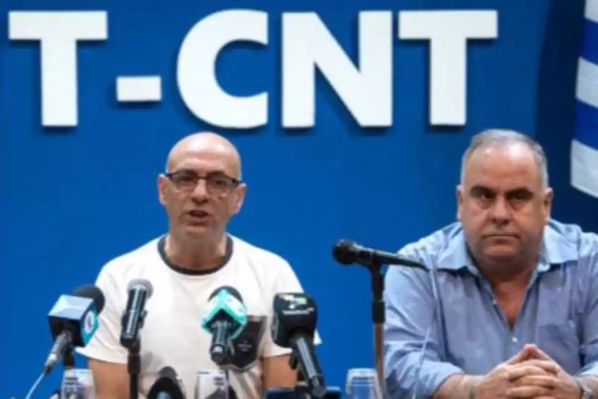 Dos hombres sentados frente a micrófonos en una conferencia de prensa con un fondo azul que tiene las letras T-CNT.