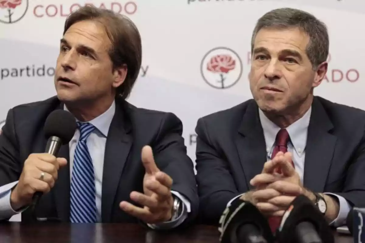 Two men in suits speaking at a press conference with microphones in front of them and a background mentioning "Colorado Party."