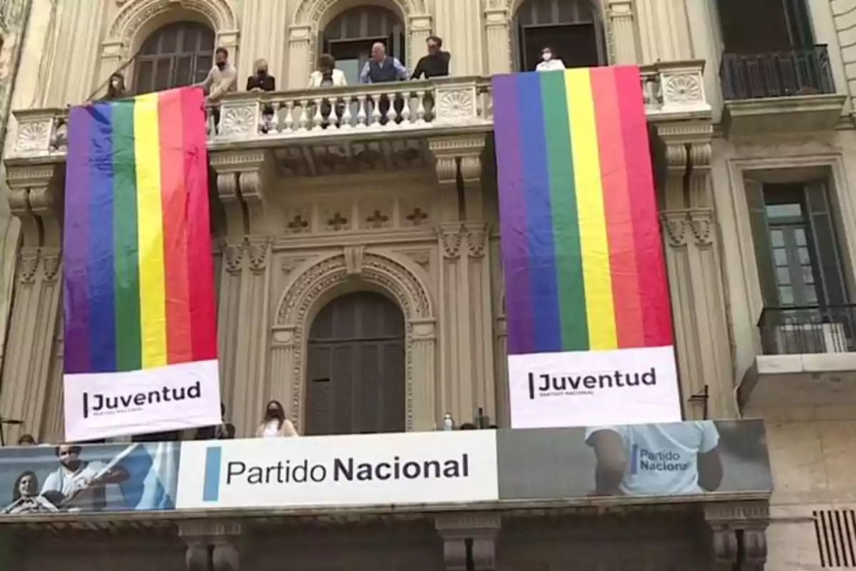 Un edificio con dos grandes banderas de arcoíris colgando desde un balcón, con personas de pie en el balcón y un cartel que dice "Juventud Partido Nacional".