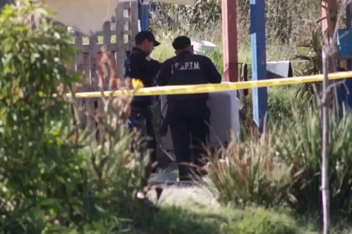 Dos personas con uniformes oscuros están de pie detrás de una cinta amarilla en un área al aire libre rodeada de vegetación.