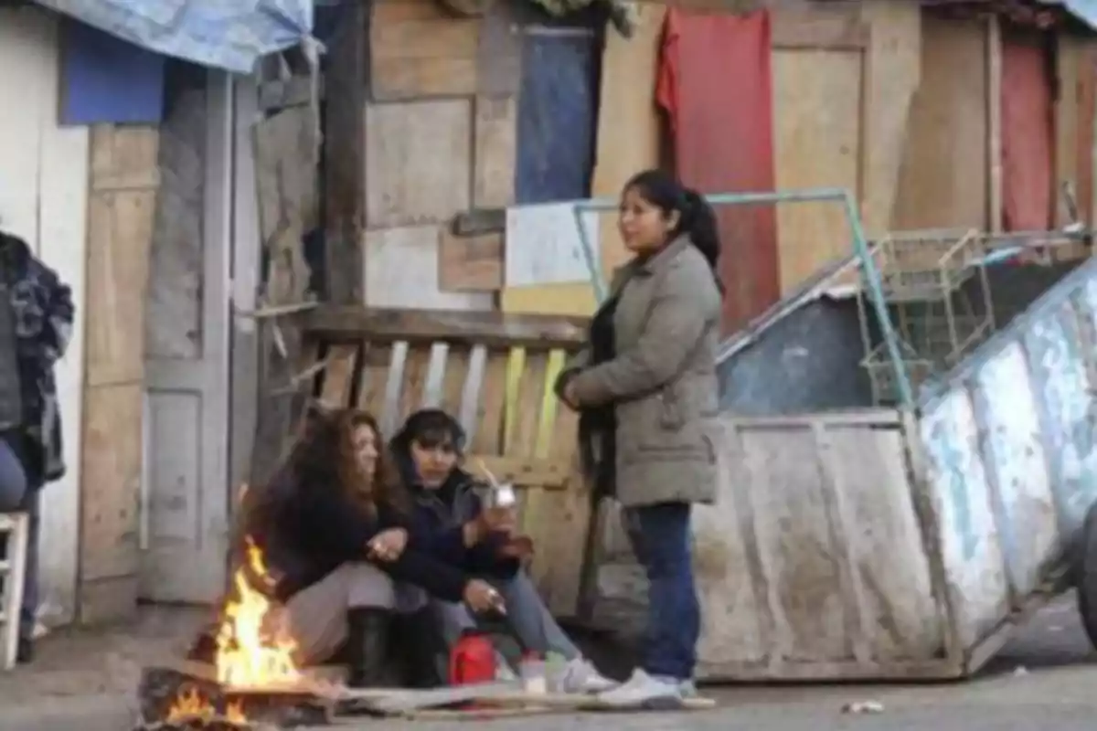 Tres personas están reunidas alrededor de un pequeño fuego frente a una estructura improvisada hecha de madera y metal.