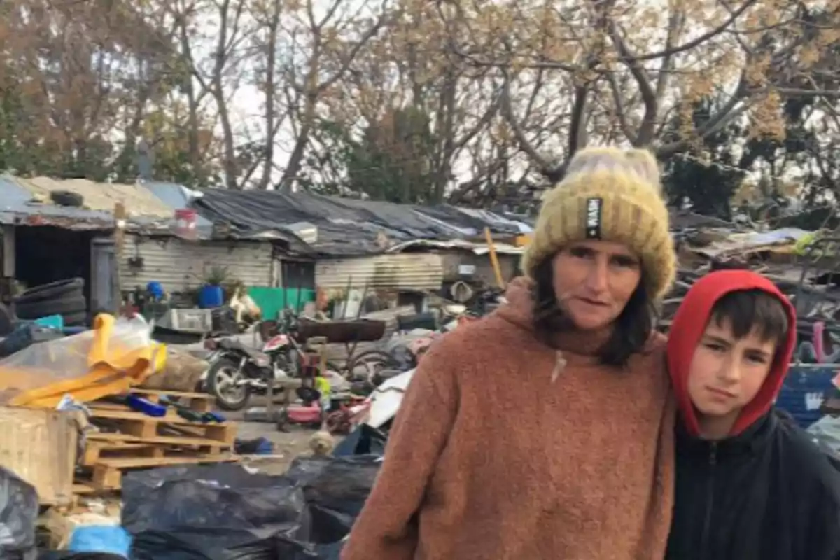 A woman and a child in warm clothing are standing in front of a cluttered environment with trees and makeshift structures in the background.