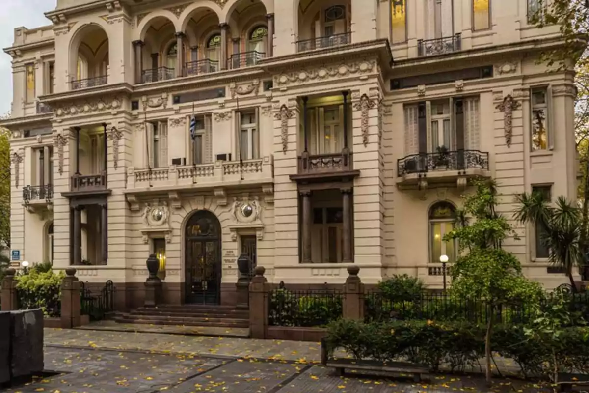 Edificio histórico de estilo neoclásico con fachada ornamentada y balcones en un entorno urbano con árboles y hojas caídas en el suelo.